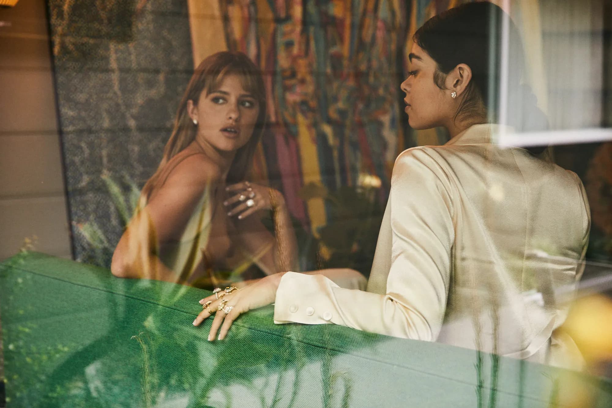 A photograph of two models behind glass sitting on a green couch wearing Heidi Gibson rings